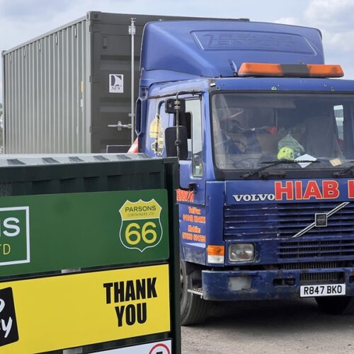 Blue HIAB lorry carrying a green 20ft container, passing the Parsons Containers signage.