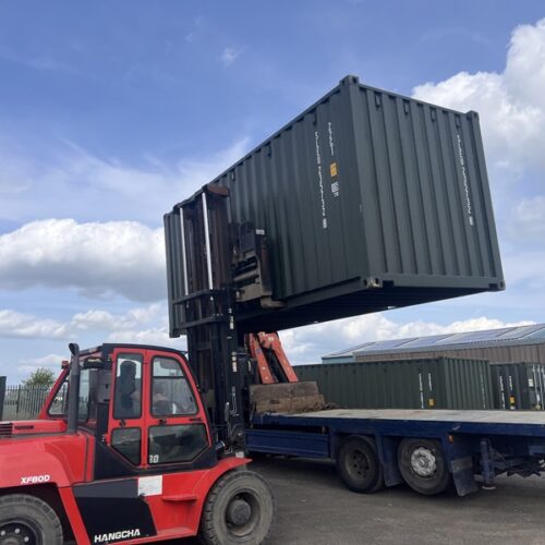 Red forklift lifting a green 20ft shipping container onto a flatbed lorry under a clear blue sky.